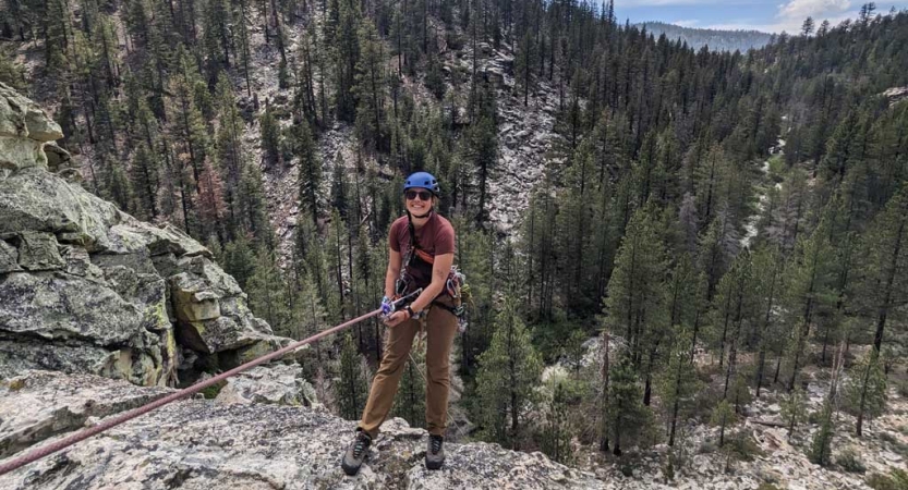 A person wearing safety gear is secured by ropes as they lean over the edge of a cliff, with trees below them. 